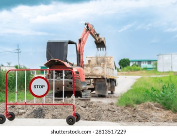 Red excavator loading soil into the back of a small dump truck at a construction site behind a temporary red stop sign barrier.