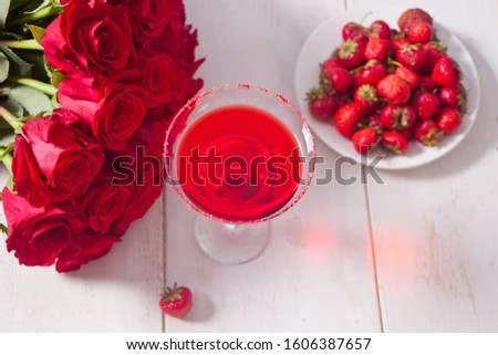 Red exotic alcoholic cocktail in clear glass, plate with fresh strawberries and red roses on the wooden white table for romantic dinner.