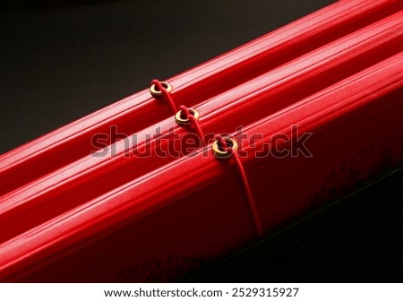 Red fabric features elegant, parallel lines secured by gold rings with red strings, set against a dark background.