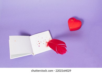 Red Feather Writing Love Letter On Clear White Paper Decorated With Hearts On Purple Background. Minimal Loving Note Idea With Heart Box. Creative Book Lover Concept.