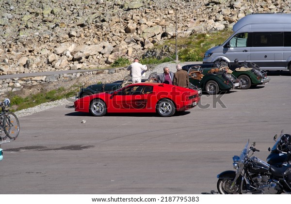 Red Ferrari sports car and dark green\
classic english sports cars at car park at Swiss mountain pass\
Sustenpass on a sunny summer day. Photo taken July 13th, 2022,\
Susten Pass, Switzerland.