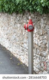 Red fire hydrant is installed on a metal pole next to a stone wall, surrounded by greenery in an urban setting