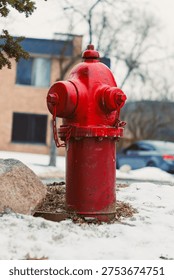A red fire hydrant is located on a snowy surface next to a building. Brown grass peeks through the snow. Nearby, a car is parked under bare trees.