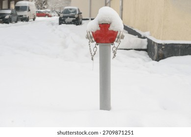 A red fire hydrant rises from deep snow in a quiet winter street, its metal caps and chains partly buried under fresh snowfall with parked cars and buildings in the background.