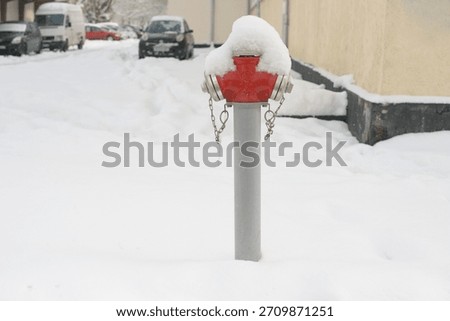 A red fire hydrant rises from deep snow in a quiet winter street, its metal caps and chains partly buried under fresh snowfall with parked cars and buildings in the background.
