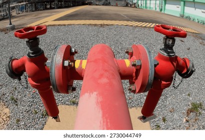 A red fire hydrant with two valves stands prominently in an industrial area. Surrounding the hydrant is gravel, with a large open space visible in the background.