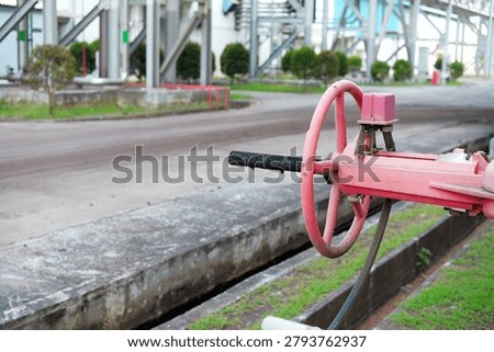 Red fire hydrant valve on industrial pipe in factory yard. Concrete edges and paved road surround the setup. Background shows industrial buildings and greenery