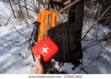 Red First Aid Kit Being Stowed Into Orange Backpack Pocket On Snowy Slope, Close Moment Of Organizing Medical Supplies, Emphasis On Compact Kit Access And Practical Packing For Winter Outing