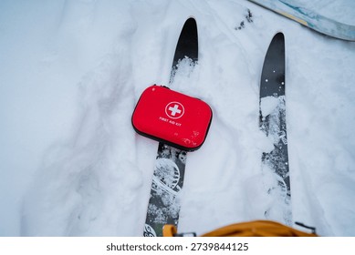 Red First Aid Kit Between Skis On Snowy Trail, Brief Rest Stop During Winter Ski Hike With Gear Staged For Quickaccess Medical Need, Calm Overcast Sky And Distant Tree Line, Practical Alpine Mood