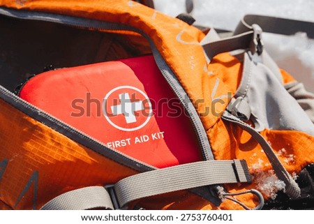 Red First Aid Kit Inside Backpack, Compact Medical Kit Nestled In Open Orange Pack, White Cross Emblem Visible, Water Droplets On Fabric, Ready For Urban Emergency Response, Paramedic Supply For Quick