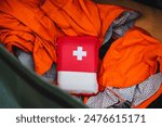 A red first aid kit with a white cross is shown from above, surrounded by bright orange safety gear. It contains essential emergency supplies for outdoor activities and camping safety