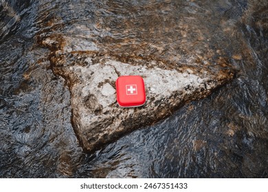 A red first aid kit with a white cross symbol on rocky surface by a stream. Essential for outdoor emergencies, survival in wilderness, and medical aid. Perfect for adventure, camping, or travel - Powered by Shutterstock