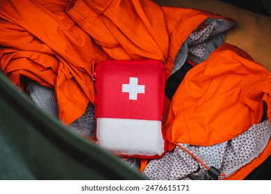 A red first aid kit with a white cross is shown from above, surrounded by bright orange safety gear. It contains essential emergency supplies for outdoor activities and camping safety