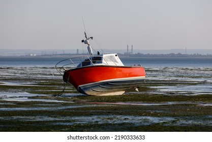 A Red Fishing Boat Lies On Its Hull At Low Tide On Mud Flats On The River Thames Estuary In Essex, England  