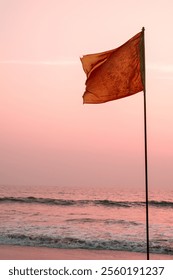 Red flag flying in the wind at the beach with sea waves and orange sky in the background at sunset in the evening. Tsunami warning, red flag waving, lifeguard rescue, windy evening.