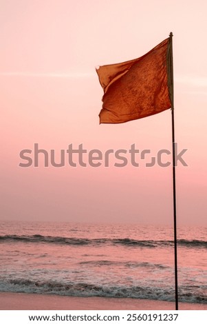 Red flag flying in the wind at the beach with sea waves and orange sky in the background at sunset in the evening. Tsunami warning, red flag waving, lifeguard rescue, windy evening.