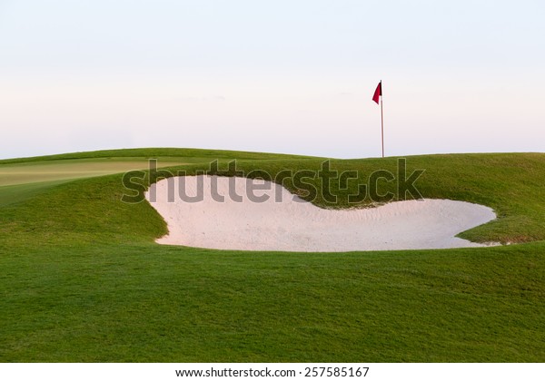 Red flag of golf hole above sand trap or bunker on\
beautiful course at sunset