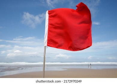Red flag on the Beach. A red flag flies in the wind on a windy but sunny summer's day, warning swimmers of dangerous surf conditions. Red flag, Cornish Beach, summer, sand, blue sky, white water 
