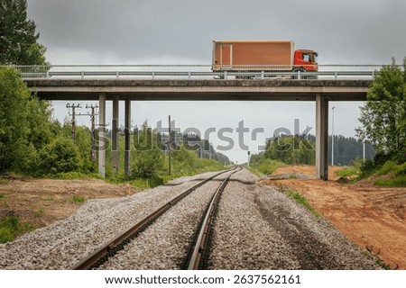 A red freight truck travels across a concrete bridge spanning railway tracks in a rural area, with power poles, gravel beds, and a forested backdrop under cloudy skies.