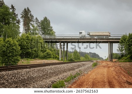 A red freight truck travels across a concrete bridge spanning railway tracks in a rural area, with power poles, gravel beds, and a forested backdrop under cloudy skies.