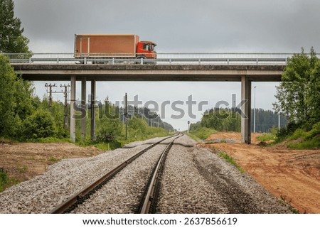 A red freight truck travels across a concrete bridge spanning railway tracks in a rural area, with power poles, gravel beds, and a forested backdrop under cloudy skies.