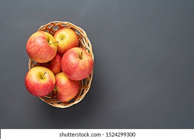 Lot Of Red Fresh Whole Ripe Apples Full Of Vitamins Lies In Wooden Wicker Basket And Old Dark Scratched Concrete Table On Kitchen. Harvest Or Diet Concept. Space For Text. Top View