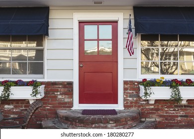 A Red Front Door Of An American Home. Also Seen Is White Siding, American Flag, Blue Awning, Flowers, And Brick Steps. 