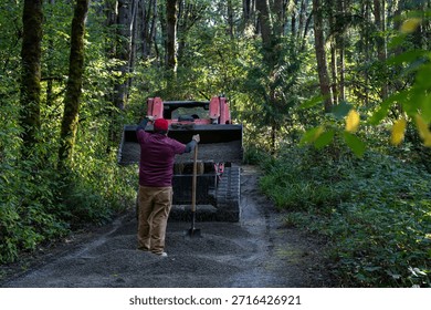 Red front loader tractor with load of fresh gravel delivering it to a forest path in public park, workman with shovel guiding the equipment operator, maintenance team hard at work
