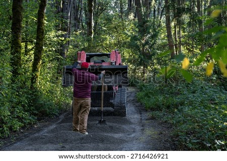 Red front loader tractor with load of fresh gravel delivering it to a forest path in public park, workman with shovel guiding the equipment operator, maintenance team hard at work

