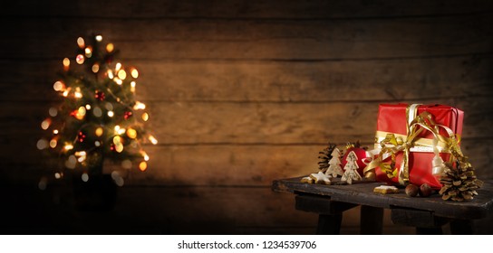 red gift box and christmas decoration on an old stool, blurry tree with lights in the background against a rustic wooden wall, copy space, panoramic format, selected focus - Powered by Shutterstock