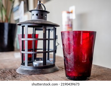 Red Glass Cup In Front, Old Lantern Frame And Another Red Cup In The Background. These Hold Tealight Candles As Modern Home Decoration Items.