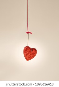Red glittery heart hooked with piece of metal hanging on pink thread on a beige background. Shoot taken from 45 degree angle. Shoot taken from Dutch angle.