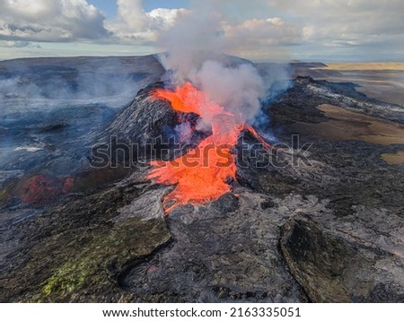Red glowing lava flows from a volcano in Iceland's Reykjanes Peninsula. View of the crater from above. Steam and smoke near the crater. cooled magma around craters. clouds in the sky at day