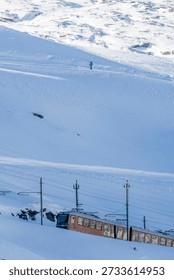A red Gornergrat train traversing through a pristine snow-covered alpine landscape. The train moves along railway tracks with electrical power lines under a clear, bright sky. A solitary skier appears in the distant background, enjoying the sunny winter day.