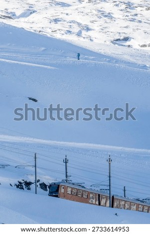 A red Gornergrat train traversing through a pristine snow-covered alpine landscape. The train moves along railway tracks with electrical power lines under a clear, bright sky. A solitary skier appears in the distant background, enjoying the sunny winter day.