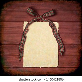 Red And Green Plaid Christmas Bow With Paper With Ragged Edges On Brown Rustic Wood Boards Background With Copy Space. The Tones Are Dark And Masculine.