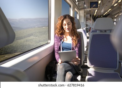 Red Hair Happy Woman Dressed In Purple And Blue, Traveling By Train Sitting Reading Or Watching Digital Tablet Or Ebook