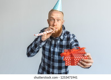 Red Hair Mature Man Wearing Birthday Cap Standing Isolated On Grey Wall Giving Gift Box To Camera Close-up Blowing Party Horn Smiling Cheerful