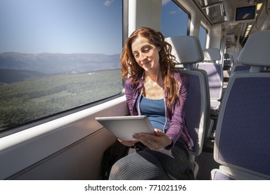 Red Hair Smiling Woman Dressed In Purple And Blue, Traveling By Train Sitting Reading Digital Tablet