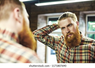 Red haired handsome guy with beard looking in mirror and enjoying his reflection