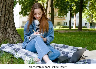 Red Haired Caucasian Woman Studying Remotely On Laptop, Writing Down Information In Notebook, Outside.Education Concept. Homework