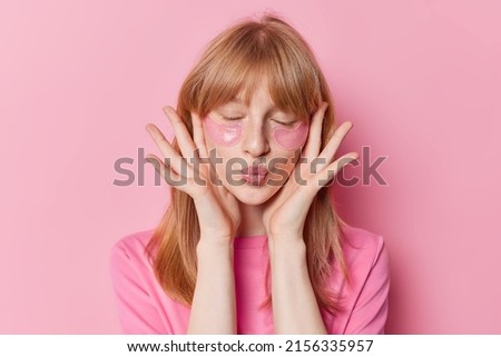 Red haired freckled girl keeps lips folded has freckled skin applies hydrogel patches under eyes undergoes beauty procedures wears casual t shirt isolated over pink background. Facial treatment