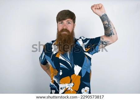 red haired man wearing printed shirt over white studio background feeling serious, strong and rebellious, raising fist up, protesting or fighting for revolution.