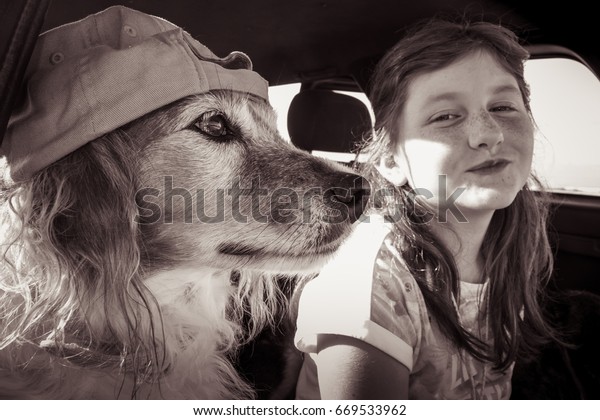 Red haired pet dog wearing baseball\
cap hanging out in a car with its red haired kid owner\
