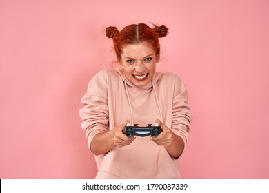 Red haired teenage girl has gamble mood plays video games with joystick on pink wall background. Female in pink hoodie has fun at home, holds playstation game controller with interest, feels crazy