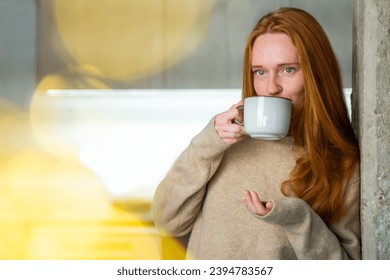 Red haired young woman wear cosy warm favorite sweater at home. Adult Drinking hot tea with herbs, while taking a break working at home. Candid moment of relaxation and tranquility. Wellness selfcare - Powered by Shutterstock