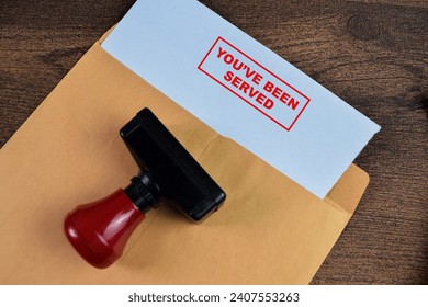 Red Handle Rubber Stamper and You've Been Served text above brown envelope isolated on wooden background