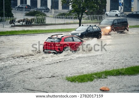 Red hatchback creates massive white splash while driving through deep flood water on city street during heavy seasonal downpour with other cars visible in the background. Cars navigating flooded road 