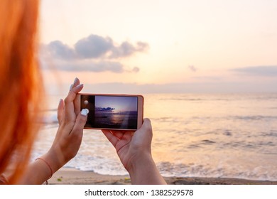 Red head girl's hand holding smart phone taking sunset photo on the beach. Mobile phone with sunset view