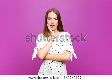 red head pretty woman open-mouthed in shock and disbelief, with hand on cheek and arm crossed, feeling stupefied and amazed against purple wall